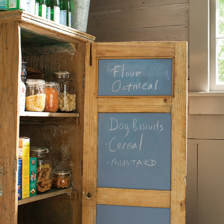 A pantry cabinet with an open door that has been painted with Benjamin Moore's chalkboard paint on the inside, with a list of food items written in chalk on the door. 